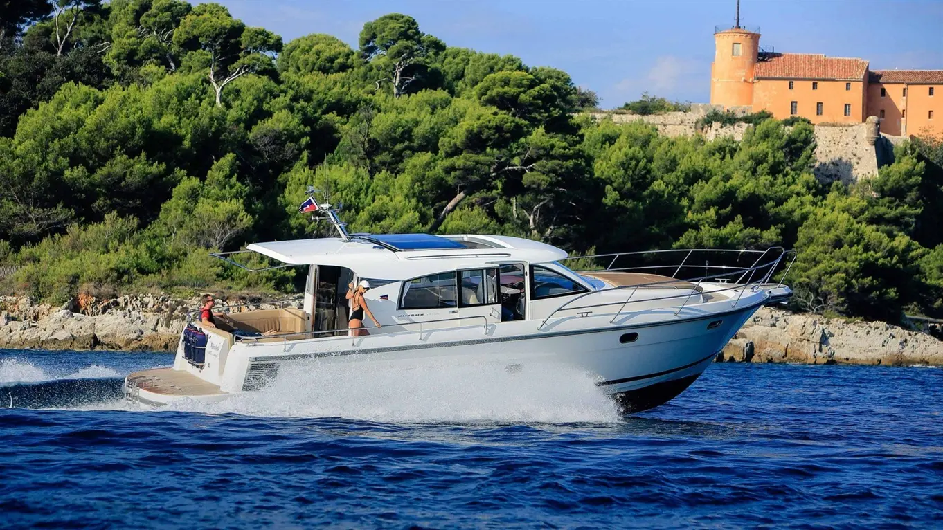 A white motorboat with two people onboard speeds through blue waters near a rocky shoreline and lush green trees.